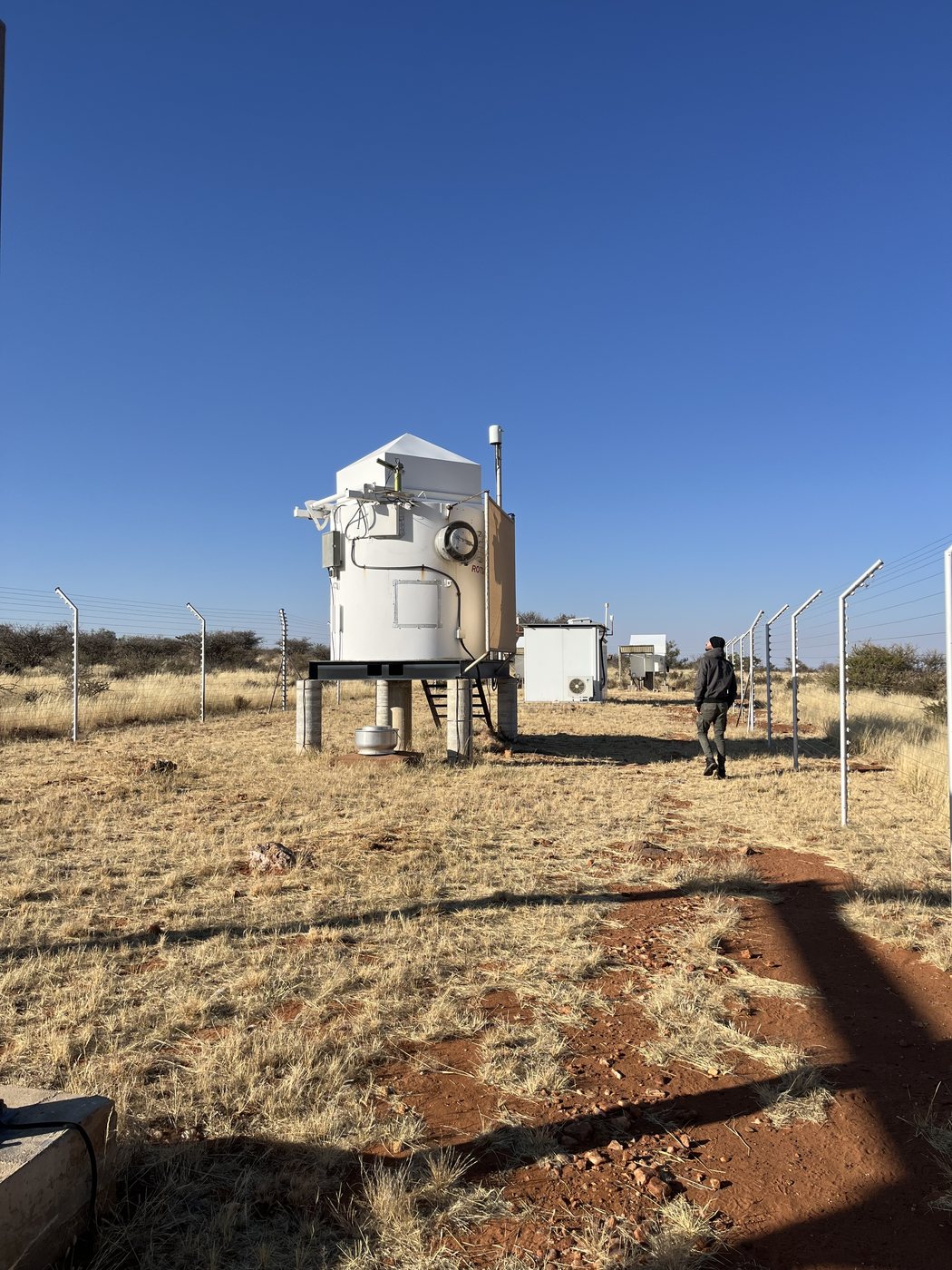 The closed ROTSE-IIIc enclosure standing on its legs inside a fenced compound in dry Namibian grassland